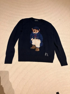Mörkblå stickad tröja Polo Bear RL - Mörkblå stickad tröja från Polo Ralph Lauren med ikoniska Polo Bear-motivet framtill. Tröjan har rund halsringning, ribbstickade muddar och RL-broderi nedtill. Perfekt för dig som gillar klassisk och lekfull stil.
