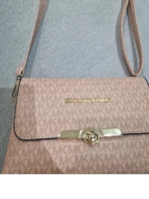 Rosa axelväska från Michael Kors - Snygg rosa axelväska från Michael Kors med guldiga detaljer och MK-logga över hela väskan. Väskan har ett lock med vridspänne i guld och justerbar axelrem. Perfekt för dig som gillar stilrena accessoarer med lyxig känsla.