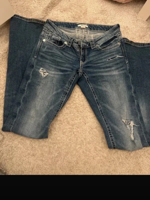 Blå bootcut jeans med slitningar - Snygga blå bootcut jeans med slitningar och ljusare tvätt på låren. Jeansen har klassisk femficksdesign, låg midja och detaljerade slitningar på benen för en trendig look. Perfekta för dig som gillar en avslappnad och cool stil.