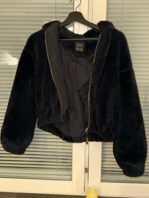 Svart teddyjacka med huva från Bershka - Mysig svart teddyjacka från Bershka med dragkedja och stor huva. Jackan är kortare i modellen och har resår i midjan samt vid ärmsluten. Perfekt för att hålla sig varm och se stylish ut under kyliga dagar.