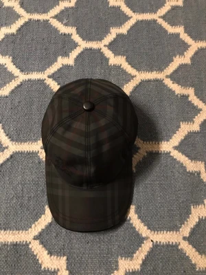 Burberry  keps  - Snygg keps från Burberry med klassiskt rutmönster i mörka färger. Justerbar rem baktill i läder med präglad Burberry-logga. Perfekt accessoar för en trendig och stilren look.