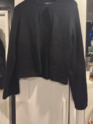 Svart kofta från Gina Tricot - Enkel och stilren svart kofta från Gina Tricot. Koftan är rak i modellen, har långa ärmar och är öppen framtill med en knapp. Mjuk och lite fluffig yta, perfekt att slänga på sig över en topp.