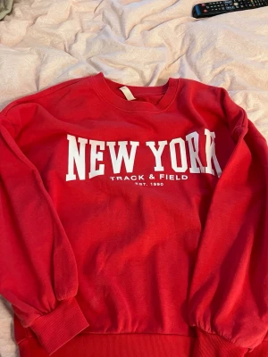 Röd sweatshirt med New York-tryck - Cool röd sweatshirt med vitt tryck där det står 'NEW YORK Track & Field Est. 1990' på bröstet. Tröjan har rund hals, ribbade muddar och är perfekt för en avslappnad streetstyle-look. Passar S, M och är lite oversize