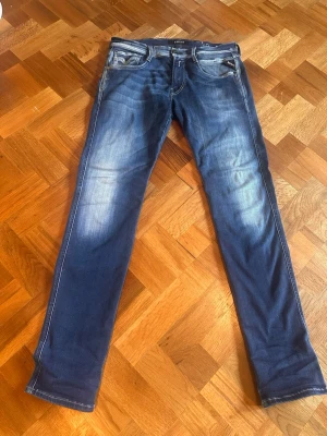 Blå anbass jeans från replay  - Säljer ett par blå slim fit jeans från replay med snygga slitningar och klassiska fem fickor. Jeansen har vita kontrastsömmar och är tillverkade i ett mjukt denimtyg som sitter skönt. Perfekta för dig som gillar en modern och avslappnad stil. W30 L32 