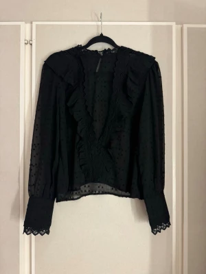 Svart blus  - Svart transparent blus med volangdetaljer längs v-ringningen och spets vid ärmslut och hals. Prickigt mönster i tyget och långa ärmar. 