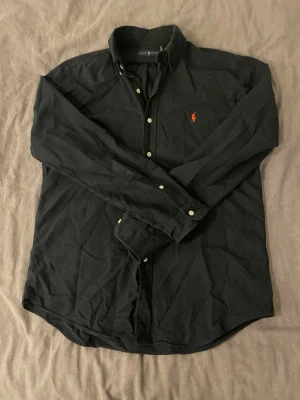 Svart skjorta från Ralph Lauren - Klassisk svart skjorta från Ralph Lauren med lång ärm och knappar framtill. Skjortan har den ikoniska röda logotypen broderad på bröstet och är tillverkad i mjuk bomull. Perfekt för en stilren och avslappnad look. Seriösa bud kan godkännas.
