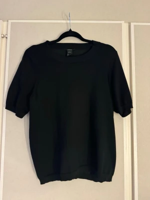 Svart stickad topp  - En svart stickad topp från Lindex med rund halsringning och korta ärmar. Enkel och stilren design som passar perfekt till jeans eller kjol. Mjuk och bekväm kvalitet med ribbade kanter vid hals, ärmslut och nederkant.