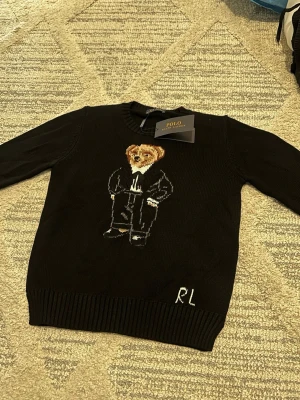 Svart stickad tröja Polo Bear RL - Svart stickad tröja från Polo Ralph Lauren med den ikoniska Polo Bear-björnen på bröstet. Tröjan har rund hals, långa ärmar och RL-broderi nedtill. Materialet är mjukt och bekvämt, perfekt för en trendig och avslappnad look. Kvitto finns med, bara för seriösa köpare tack ☺️ 