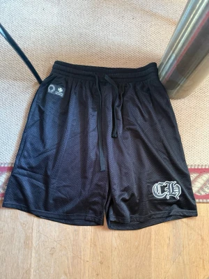 Svarta meshshorts från Chrome Hearts - Svarta shorts från Chrome Hearts i meshmaterial med snörning i midjan och vit logga tryckt på ena benet. Perfekta för en avslappnad och sportig stil. Kommer i originalpåse med tydlig Chrome Hearts-logga.