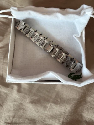 Silverfärgat armband från Lacoste - Snyggt armband i silverfärgad metall från Lacoste. Länkarna har ett graverat mönster som ger en cool och modern look. Kommer i original ask med klassiska Lacoste-loggan på locket. Perfekt accessoar för dig som gillar stilrena detaljer.