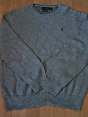 Grå tröja från Polo Ralph Lauren - Klassisk grå stickad tröja från Polo Ralph Lauren med rund halsringning och diskret broderad logga på bröstet. Tröjan har ribbade muddar vid ärmslut och nederkant, perfekt för lager på lager under höst och vinter. Axelbredd: 46cm / Längd: 61cm. Nypris: 2295 / Mitt pris: 449kr