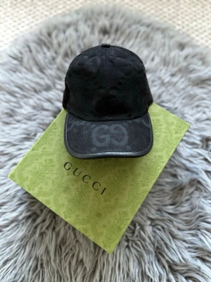 Svart keps från Gucci med GG-mönster - Snygg svart keps från Gucci med det klassiska GG-mönstret ton-i-ton över hela kepsen. Justerbar rem baktill för perfekt passform och böjd skärm. Tillverkad i ett slitstarkt tyg som ger en exklusiv känsla. Perfekt accessoar för en trendig streetwear-look.
