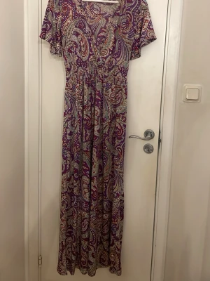 Långklänning med paisleymönster från Elissa - Långklänning från Elissa med färgstarkt paisleymönster i lila, rött och beige. Klänningen har v-ringning, korta ärmar och ett luftigt fall. Perfekt för dig som gillar bohemisk stil och vill sticka ut med mönster.