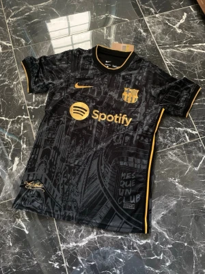 Svart FC Barcelona matchtröja Nike - Snygg svart FC Barcelona matchtröja från Nike med guldfärgade detaljer och loggor. Tröjan har tryck av Spotify och klubbmärket på bröstet samt diskreta mönster i tyget. Tillverkad i lätt och ventilerande material, perfekt för fotboll eller träning.