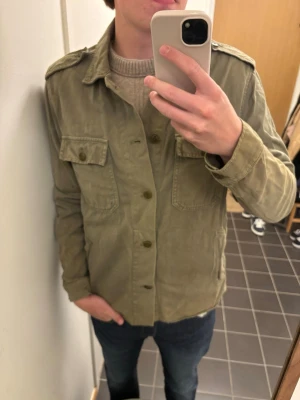 Olivgrön overshirt från Zara Man - Snygg olivgrön overshirt från Zara Man i storlek L. Klassisk design med två bröstfickor med lock och knappar framtill. Perfekt lager-på-lager-plagg med avslappnad passform och axelklaffar. Tillverkad i mjukt bomullstyg.