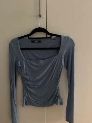 Ljusblå långärmad topp från BikBok - Supersnygg ljusblå långärmad topp från BikBok med fyrkantig ringning och draperad effekt framtill. Toppens material känns mjukt och stretchigt, perfekt för en trendig look. Passar dig som gillar stilrena och moderna plagg.