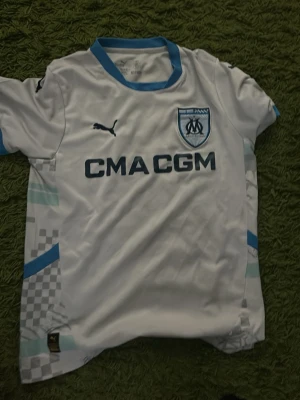 Marseille matchtröja Puma vit/blå - Säljer en vit Marseille matchtröja från Puma med blå detaljer och klubbmärke på bröstet. Tröjan har trycket 'CMA CGM' på framsidan och ljusblå inslag vid krage och ärmslut. Perfekt för dig som älskar fotboll och vill stötta ditt lag.