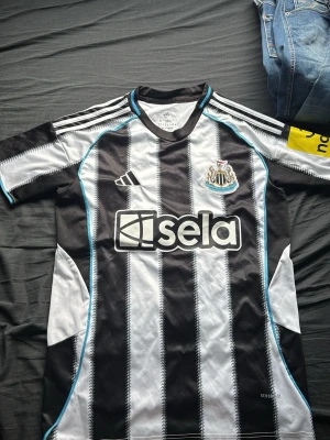 Newcastle United matchtröja Isak 14 - Snygg Newcastle United matchtröja med svarta och vita ränder, klubbmärke på bröstet och sponsorlogga på framsidan. Adidas-logga och gula detaljer på ärmen. På ryggen står det ISAK och nummer 14. Tillverkad i lätt och ventilerande material. Storlek M men kan även passa S