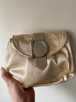 Beige väska/necessär Dior - Snygg beige väska/necessär från Dior beauty/parfyms med quiltat lock och stor silverfärgad D-detalj framtill. Necessär är gjord i glansigt satinliknande material och har en klassisk, rundad form. Perfekt för dig som gillar lyxiga accessoarer med ikonisk design.