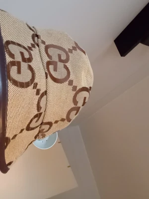 Beige bucket hat från Gucci - Säljer en beige bucket hat från Gucci med det klassiska GG-mönstret i brunt. Hatten har en rund form och är tillverkad i canvasliknande material med mörkbrun kant runt brättet. Perfekt accessoar för en trendig look.
