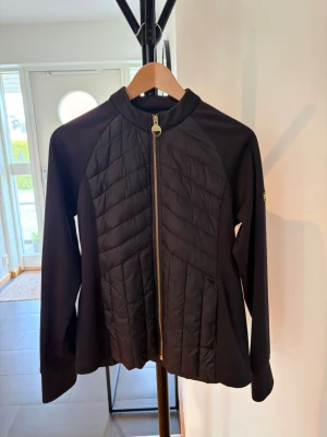 Svart hybridjacka med quiltade paneler - Snygg svart jacka med quiltade paneler fram och bak, hel dragkedja i guld och ståkrage. Jackan har långa ärmar och en sportig look, perfekt för lager-på-lager. Materialet är en mix av slät textil och vadderade partier. Liten logga på ärmen.
