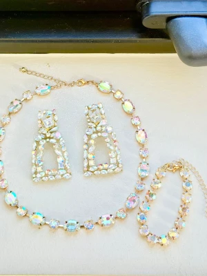 Set med gnistrande smycken  - Säljer ett matchande smyckeset med halsband, armband och örhängen med stenen aurora boreale. Infattade i guldfärg kedjor. Alla delar är dekorerade med stora, skimrande, iriserande stenar i olika former som fångar ljuset. Örhängena har en unik, rektangulär design och kedjorna är justerbara på både armband och halsband. Perfekt till studentbal, fest, bröllop mm. Ej använt! 
