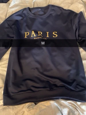 Svart sweatshirt med Paris-tryck - Cool svart sweatshirt med gul text 'PARIS Romantic City' på bröstet. Tröjan har rund halsringning och långa ärmar. Perfekt för dig som gillar enkel men snygg streetstyle. Materialet känns mjukt och bekvämt mot huden.