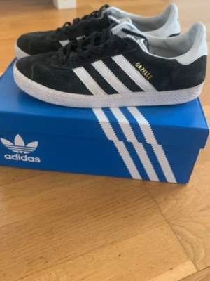 Adidas Gazelle svarta sneakers - Svarta Adidas Gazelle sneakers i mocka med vita detaljer och klassiska tre ränder på sidorna. Låg modell med vit sula och Adidas-logga på hälen. Snygg och tidlös design som passar till det mesta.
