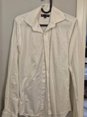 Vit slim fit skjorta från Dressmann - Klassisk vit skjorta från Dressmann i slim fit-modell. Skjortan har lång ärm, knappar framtill och en bred krage. Tillverkad i bomull för en skön känsla och stilren look. Perfekt för dig som gillar en clean och enkel stil.