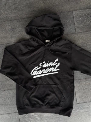 Svart Saint Laurent hoodie - Snygg svart hoodie från Saint Laurent med vit logga i graffiti-stil på bröstet. Klassisk känguruficka framtill och justerbar huva med snören. Tillverkad i mjukt bomullsmaterial som känns skönt mot huden. Perfekt för en avslappnad och trendig look.