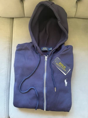 Marinblå hoodie från Polo Ralph Lauren - Snygg marinblå hoodie från Polo Ralph Lauren med dragkedja framtill, huva med snörning och klassisk logga broderad på bröstet. Tillverkad i mjuk bomull och har två fickor framtill. Perfekt för en chill och stilren look.