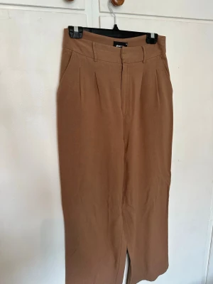Bruna vida kostymbyxor från Gina Tricot - Snygga bruna kostymbyxor från Gina Tricot med hög midja och vida ben. Byxorna har pressveck framtill, hällor för skärp och stängs med dragkedja och knapp. Säljes då dom är för stora 🌸