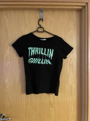 Svart t-shirt med tryck från H&M - Svart t-shirt från H&M med coolt grönt tryck där det står 'Thrillin Chillin' på framsidan. Klassisk passform med rund hals och korta ärmar. Perfekt för dig som vill ha en enkel men ändå uttrycksfull look.