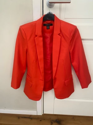 Röd orange kavaj från Amisu - Snygg orange röd kavaj från Amisu med klassisk krage och en knapp framtill. Kavajen har två fickor med lock och är helfodrad. Perfekt för att lyfta din outfit med en färgklick. Rak passform och stilren design.