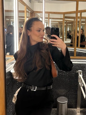 Svart långärmad satinblus med hög krage - Stilren svart blus med lång ärm och hög krage. Blusen har en rak passform och är tillverkad i ett mjukt, lätt material som ger en elegant look. Perfekt att matcha med både jeans och kostymbyxor. Inga synliga loggor eller mönster.
