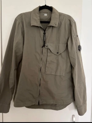 Olivgrön overshirt från C.P. Company - Säljer en olivgrön overshirt från C.P. Company med dragkedja, klassisk krage och stor bröstficka med lock. Ärmen har en ikonisk linsdetalj och extra ficka. Tillverkad i lätt bomullsmaterial, perfekt för lager-på-lager-stil.