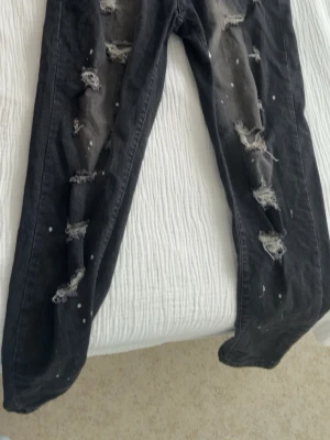 Svarta jeans med håll  - Säljer ett par svarta jeans med slitningar och hål på benen samt vita färgstänk. Jeansen har en rak passform och är i denim. Perfekta för en edgy streetstil.