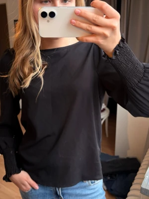 Svart blus med smockade ärmslut - Stilren svart blus med rund halsringning och långa ärmar. Ärmsluten har smockdetaljer och volangkant som ger en trendig touch. Blusen är tillverkad i ett tunt, mjukt tyg som faller snyggt.