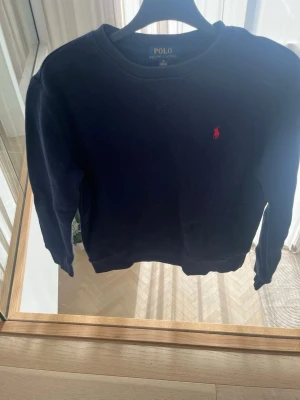 Mörkblå sweatshirt från Polo Ralph Lauren - Klassisk mörkblå sweatshirt från Polo Ralph Lauren med rund halsringning och röd broderad logga på bröstet. Tröjan har långa ärmar och ribbade muddar vid ärmslut och nederkant. Perfekt för en avslappnad och stilren look.