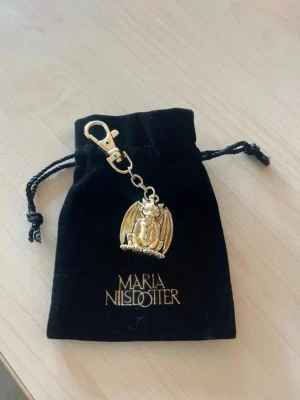 Guldfärgad draknyckelring Maria Nilsdotter - Cool nyckelring i guldfärgad metall med en detaljerad drake som hänge. Kommer med svart sammetsfodral med Maria Nilsdotter-logga. Perfekt accessoar för dig som gillar unika detaljer och fantasyvibbar.
