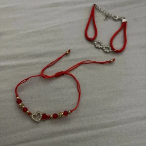 Röda armband med pärlor och hjärta - Två snygga armband i röd tråd, ett med pärlor i rött och guld samt ett hjärthänge med strass, och ett med silverfärgad detalj och justerbart spänne. Perfekta accessoarer för att lyfta din stil med färg och bling.