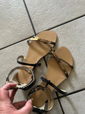 Svarta sandaler med gulddetaljer storlek 37 - Snygga sandaler med svarta remmar i lack och guldfärgade metalldetaljer. Öppen tå och justerbar rem runt vristen. Platt sula i beige och stilren design som passar till många outfits.