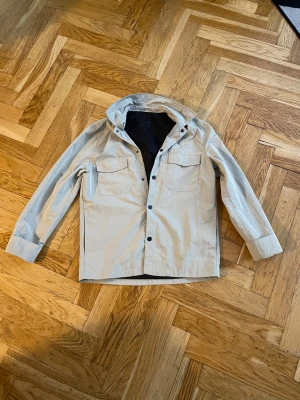 Beige overshirt från Elvine - Stilren overshirt vindjacka från Elvine med huva och svarta tryckknappar. Jackan har två bröstfickor med lock och raka ärmar. Den är i bra skick. Extra knappar medföljer vid köp . Passar till de mesta