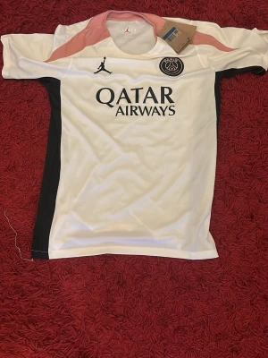 PSG x Jordan vit matchtröja - Snygg vit Paris Saint-Germain matchtröja från Jordan med svarta och rosa detaljer. Tröjan har PSG-logga på bröstet, Jordan-logga och Qatar Airways-tryck framtill. Tillverkad i lätt och ventilerande material, perfekt för fotboll eller träning.