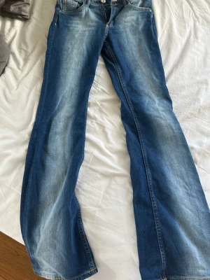 Blå bootcut jeans med slitningar - Snygga blå bootcut jeans med ljusa slitningar längs benen och klassisk femficksdesign. Jeansen har låg midja och är tillverkade i denim med en lätt stretch för skön passform. Perfekta för en avslappnad och trendig look.
