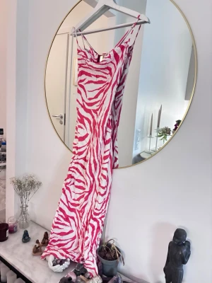 Zebramönstrad långklänning rosa/vit - Säljer en långklänning med smala axelband och ett coolt zebramönster i rosa och vitt. Klänningen har en figurnära passform och är tillverkad i ett lätt, mjukt material som känns skönt mot huden. Perfekt för dig som vill sticka ut med en unik look.