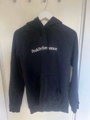 Svart hoodie från Peak Performance - Svart hoodie från Peak Performance med vit logga på bröstet. Klassisk modell med huva och dragsko samt stor magficka framtill. Mjuk bomullsblandning och ribbade muddar vid ärmslut och nederkant. Perfekt för en avslappnad stil.