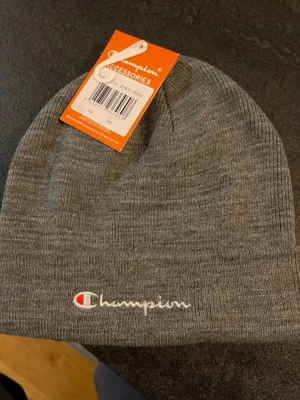 Helt ny Grå mössa från Champion one size - Snygg grå mössa från Champion med broderad logga framtill i vitt och rött. Klassisk ribbstickad design som passar perfekt till streetwear-stilen. Materialet är mjukt och stretchigt för skön passform.