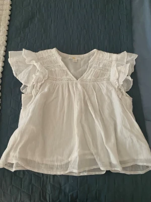 Vit volangblus med v-ringning - Söt vit blus med volangdetaljer på axlarna och v-ringad hals. Blusen har korta ärmar och är tillverkad i ett lätt, luftigt material som ger en avslappnad känsla. Perfekt för en fräsch och stilren look.