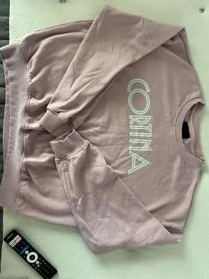 Rosa sweatshirt med tryck - Säljer en oversized ljuslila sweatshirt med trycket 'CORTINA' i vitt framtill. Tröjan har rund hals, ribbade muddar och är tillverkad i mjuk bomullsblandning. Perfekt för en avslappnad och trendig look. Står ingen storlek på den men skulle säga S